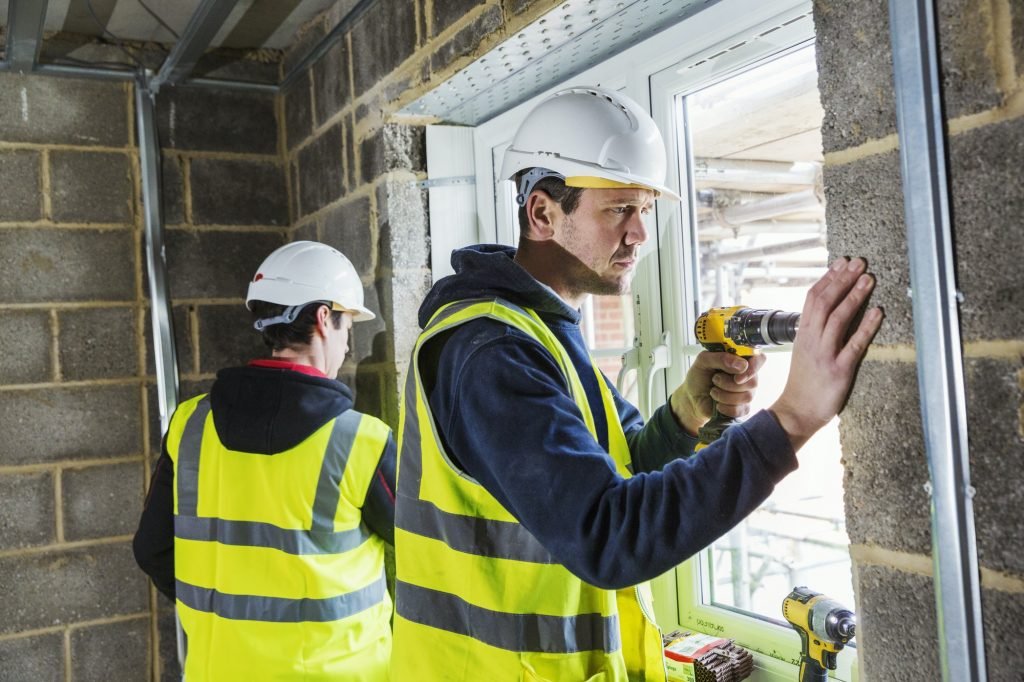 Two workmen on a construction site, builder in hard hat using an electric drill on a window frame.